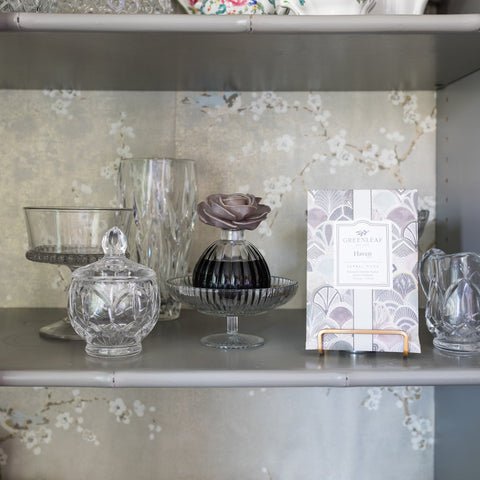 A display shelf with glassware and a floral backdrop features a crystal jar, a decorative perfume bottle with a rose cap, and a patterned box labeled Greenleaf. Large sachets from Greenleaf subtly spread their Haven fragrance, adding enchantment to the scene.