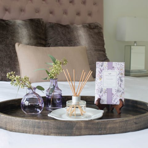 A wooden tray on the bed holds two small purple vases with greenery, a Greenleaf fragrance box, and a reed diffuser. Lavender sachets from Greenleaf complete the serene setting. The background features beige pillows and a tufted headboard.