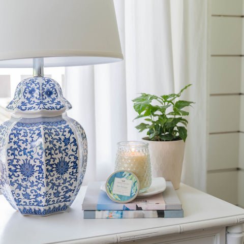 A blue and white patterned ceramic lamp sits on a white table alongside a lit Greenleaf - Spa Springs Signature Candle in a glass jar, a small potted plant, and stacked books. Sheer white curtains hang behind, enhancing the peaceful Spa Springs atmosphere.