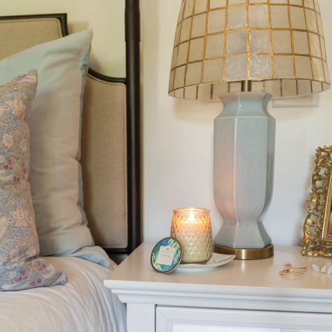 A cozy bedroom scene features a soft pillow on the bed, a decorative lamp with a checkered shade on the white nightstand, and a lit Greenleaf Spa Springs Signature Candle. A small round tin, framed photo, and Greenleaf rings add charm to the setting.