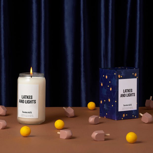 A Homesick Latkes and Lights natural soy wax candle sits on a table next to its box, surrounded by small yellow balls and wooden dreidels on a brown surface with a dark blue velvet curtain backdrop, celebrating the Festival of Lights.