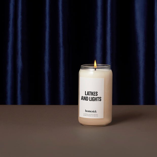 A lit Homesick candle labeled Latkes and Lights, made from natural soy wax, sits on a brown surface against a dark blue velvet curtain.