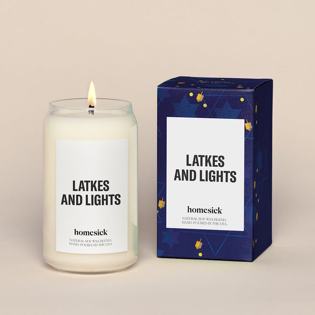 A lit Homesick Latkes and Lights candle sits beside its festive blue packaging with stars. This menorah-inspired candle is hand-poured in the USA using a natural soy wax blend for a cozy holiday atmosphere.