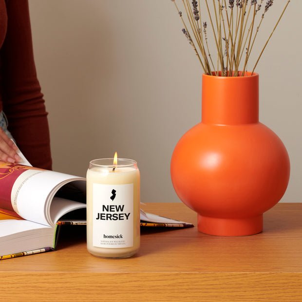 A Homesick - New Jersey Candle made from natural soy wax and premium cotton wicks is on a wooden table beside an open book and a round orange vase filled with lavender stems. A person in a rust-colored top is partially visible, leafing through the book.