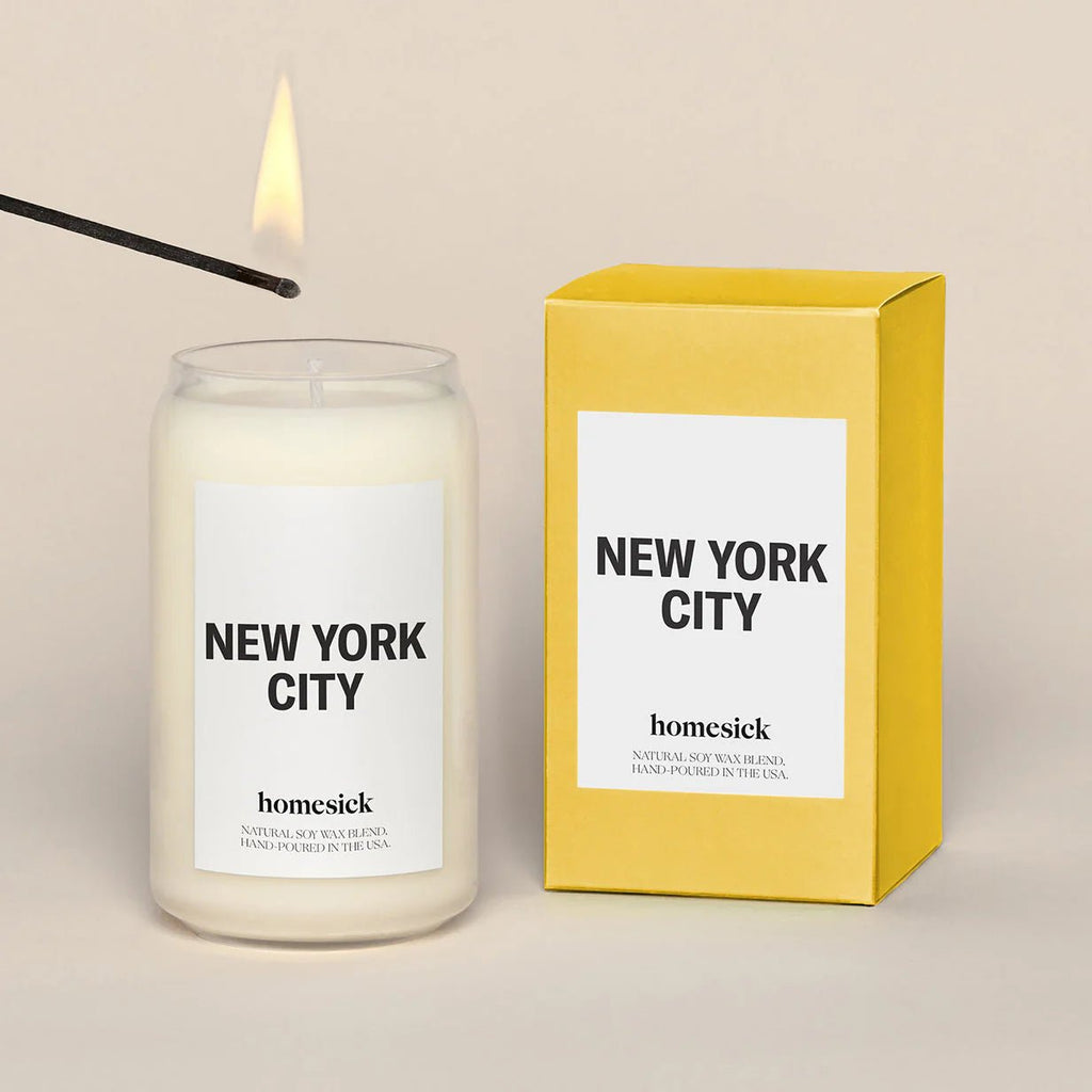 The Homesick - New York Candle flickers softly beside its vibrant yellow box, releasing a Central Park-inspired aroma. A lit match enlivens this Homesick candle, casting a city-like warmth against the stark backdrop reminiscent of NYC’s concrete jungle.