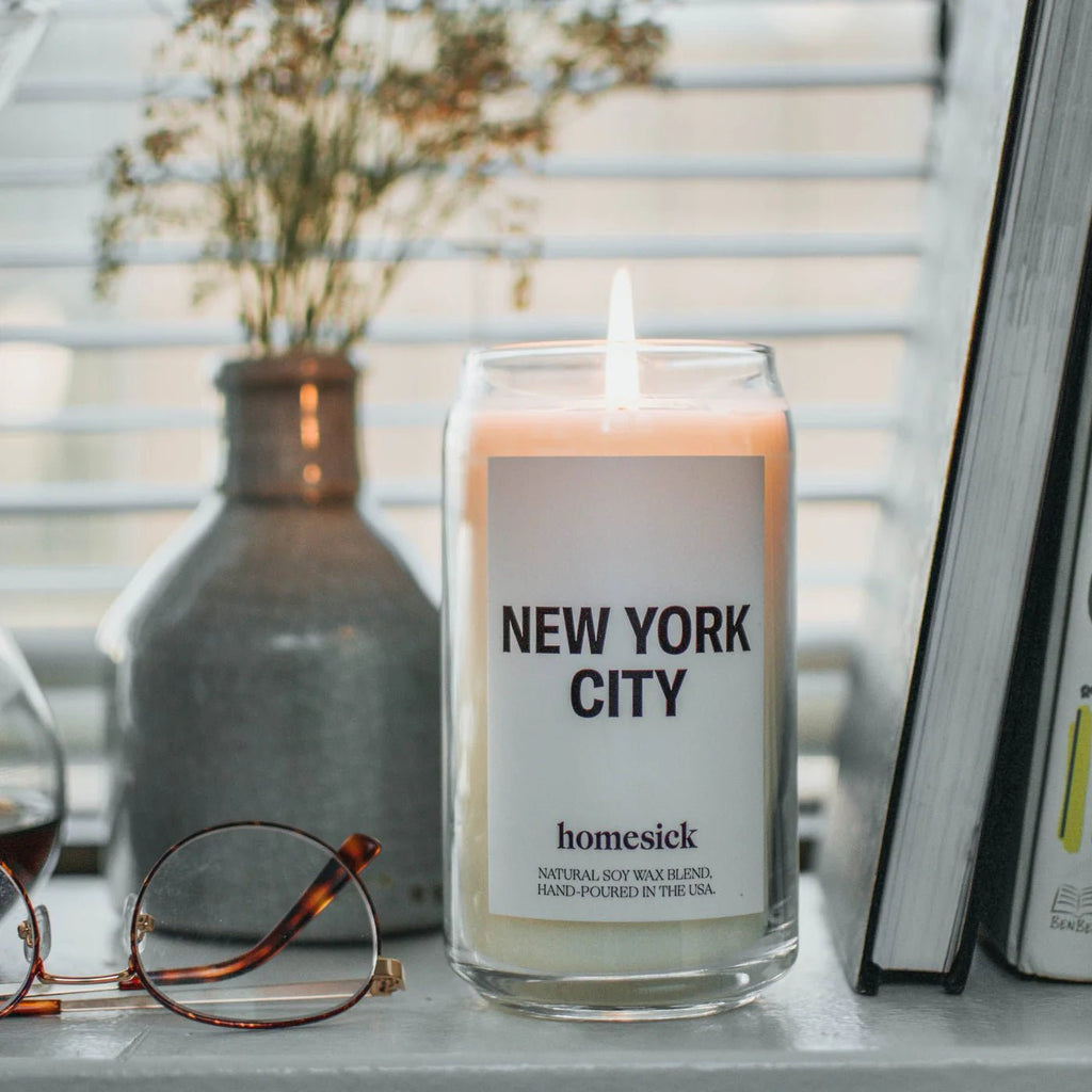 A Homesick - New York Candle by Homesick glows on a windowsill in a glass jar. Nearby, glasses rest beside a vase with dried flowers and partially visible books. Soft light filters through blinds, evoking Central Parks calm feel amid NYCs concrete setting.