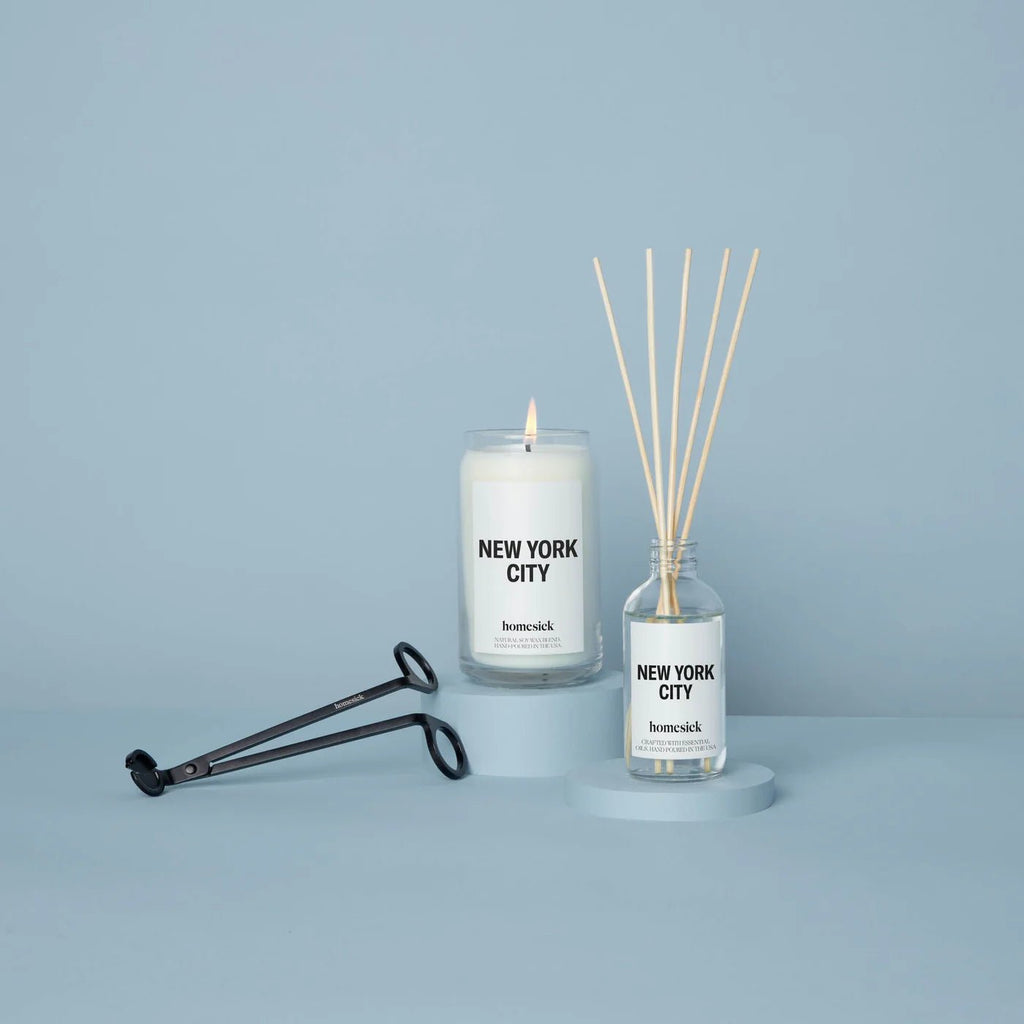 A Homesick New York Candle captures Central Parks essence beside a matching reed diffuser on a soft blue backdrop, with a black wick trimmer artfully placed diagonally, completing the urban oasis ambiance.