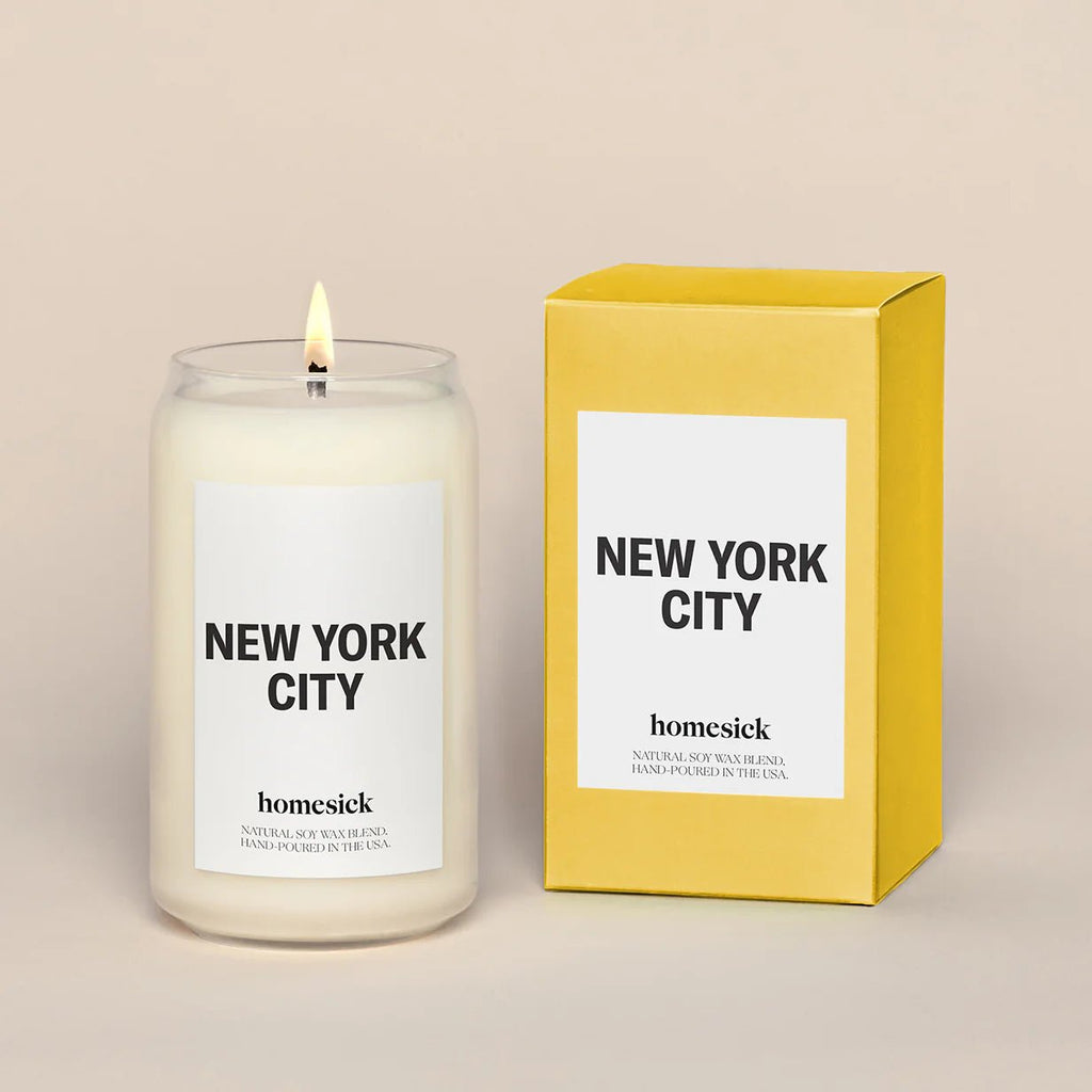 A lit Homesick - New York Candle in a glass jar sits beside its yellow box, both displaying the Homesick logo, capturing the essence of Central Park in soothing wax.