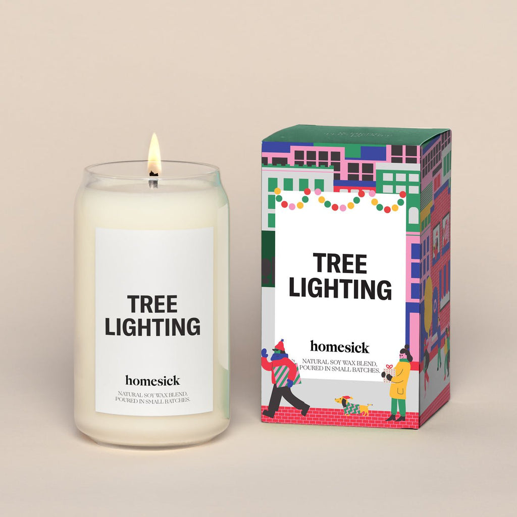 A Homesick Tree Lighting candle, crafted from natural soy wax, sits beside its colorful box adorned with festive illustrations of people celebrating. The box hints at the aromas of red currants and sugar plums.