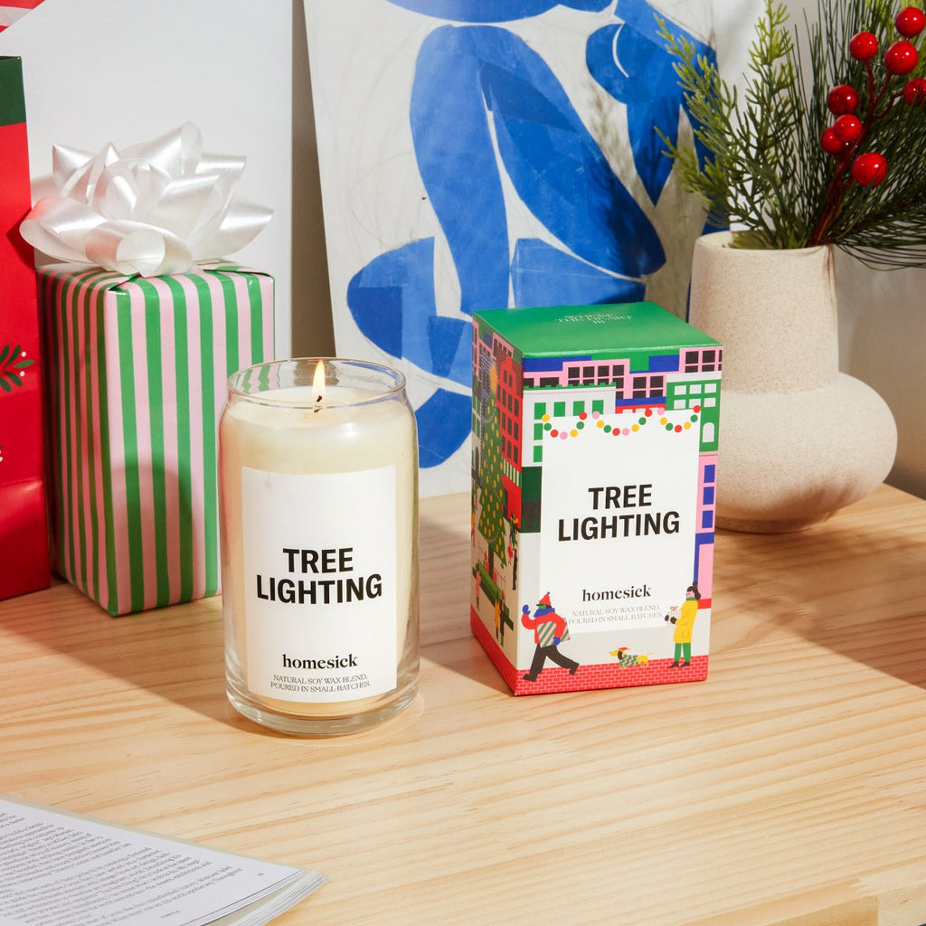 A Tree Lighting Homesick candle, crafted from natural soy wax, rests on a wooden table next to its vibrant festive box. Wrapped gifts and a potted plant with red berries appear in the background, along with a blue abstract art piece.