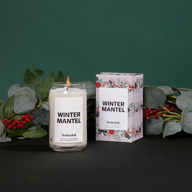 Against a dark green backdrop, a lit Homesick - Winter Mantel Candle sits next to its floral-decorated box. Crafted from soothing soy wax, it is surrounded by eucalyptus leaves and red berries for a festive touch.