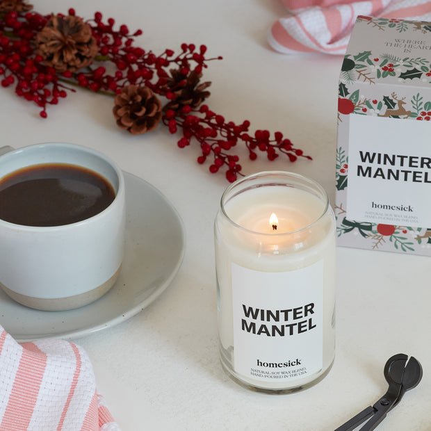 The Homesick Winter Mantel candle, made from a soy wax blend, sits beside coffee, red berries, pine cones, a matching box, pink-striped towels, and a wick cutter.