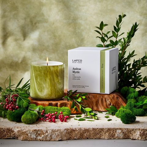 A Lafco Andean Myrtle Classic 15.5oz candle in a marbled holder sits on a wooden slab with branches, red berries, and moss, releasing an earthy Palo Santo scent. Beside it is a white Lafco New York Andean Myrtle box, against a soft green background.