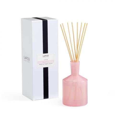 A pink glass Lafco - Blush Rose 6oz Reed Diffuser with wooden reeds stands next to its white box featuring a black stripe and the brand Lafco, offering elegant bergamot and damask rose notes.