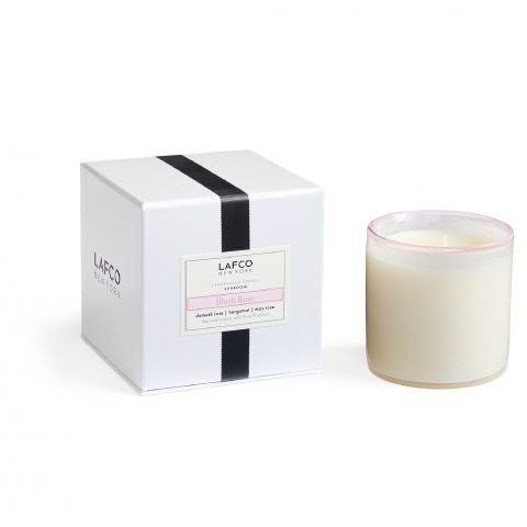 A hand-blown white glass candle sits beside a white box labeled Lafco - Blush Rose Classic 6.5 Candle. The packaging features black and pink details, with notes of Asian Pear, Bergamot, Ivy, complemented by hints of damask rose for elegance.