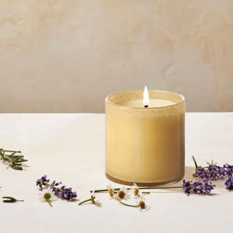 A Lafco Chamomile Lavender Classic 6.5 Candle made from soy wax flickers in a glass holder on a white surface, surrounded by scattered lavender and chamomile flowers, creating a serene atmosphere against a neutral background.