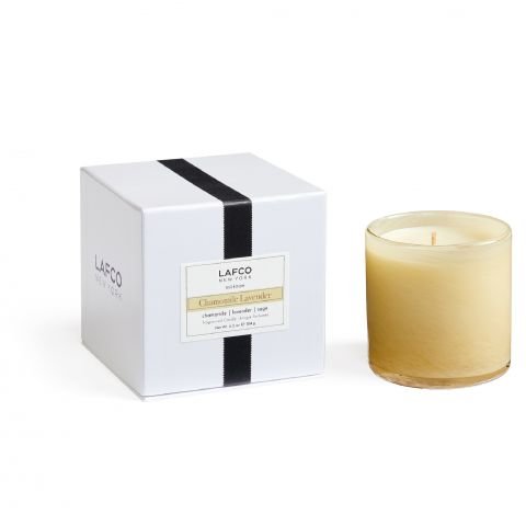 The Lafco Chamomile Lavender Classic 6.5 Candle, in a beige glass container, sits beside a white box with a black stripe labeled Lafco New York. Both the candle and the box showcase an elegant design.