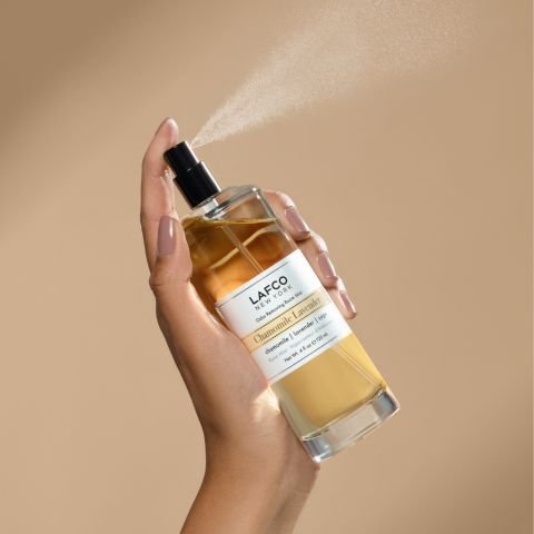 A hand sprays a labeled Lafco - Chamomile Lavender Room Mist against a beige backdrop, releasing visible chamomile-infused mist from the nozzle while the partially full bottle shows light amber liquid.