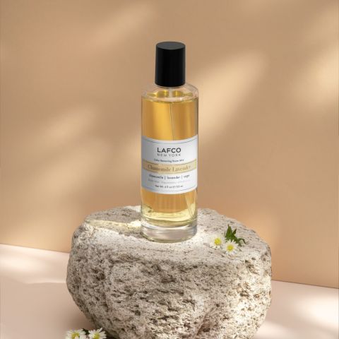 A glass bottle of Lafcos Chamomile Lavender Room Mist sits on textured stone. The lavender-infused golden liquid, capped in black, is adorned with small white daisies against a beige background.