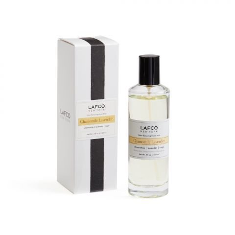 A bottle of Lafco - Chamomile Lavender Room Mist, infused with calming lavender, sits beside a sleek box. The clear bottle has a black cap, and the box features a stylish design with a white background and subtle black and yellow accents.