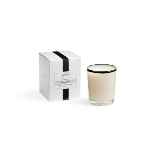 A Lafco Champagne votive candle rests in its glass holder beside a matching box, featuring a white design with black stripes and silver accents. Made from soy wax with a black-rimmed holder, the candle exudes an elegant citrus aroma.