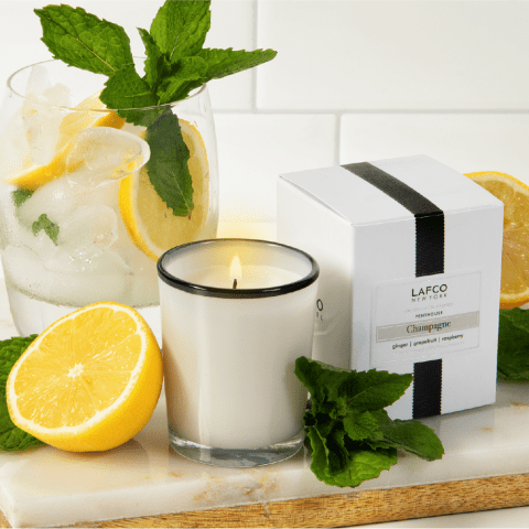 On a marble surface, a Lafco - Champagne 1.9oz Votive Candle in a glass holder sits beside its white box. The room is filled with citrus fragrance, enhanced by an iced drink with mint and lemon slices, alongside whole lemons nearby.