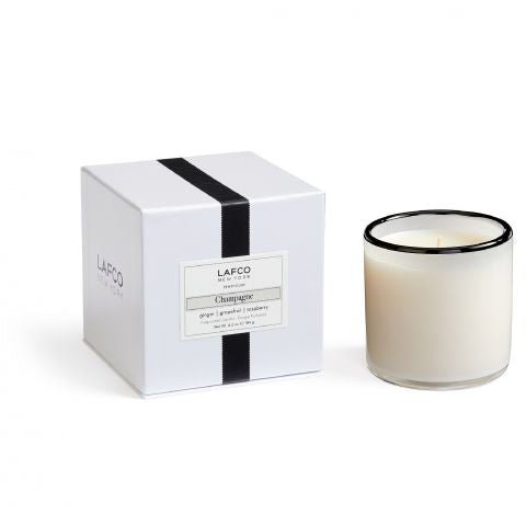 The Lafco - Champagne Classic 6.5 Candle features white soy wax in hand-blown glass with a black rim, displayed beside its white box packaging with a black stripe and Champagne label.