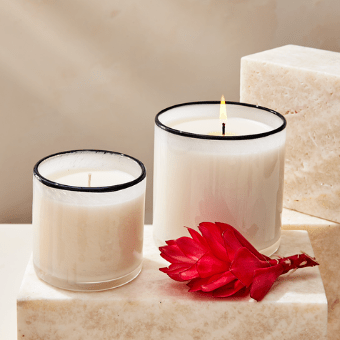 Two Lafco - Champagne Classic 6.5 Candles, in clear handblown glass holders, rest on marble blocks—one aglow, one unlit—with a vibrant red flower beside them.
