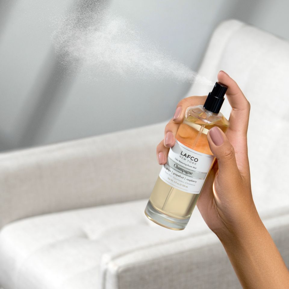A hand sprays Lafco - Champagne Room Mist from a clear bottle, filling a bright room and light-colored armchair with an elegant, luxurious scent.