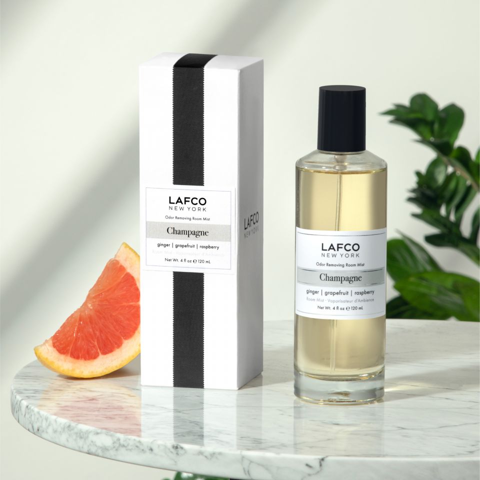 A bottle and box of Lafco - Champagne Room Mist by Lafco are displayed on marble with a grapefruit wedge nearby and green leaves in the background.