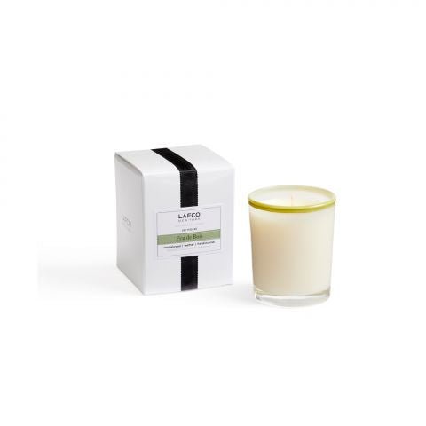 The Lafco Feu De Bois 1.9oz votive candle, exuding a subtle sandalwood scent, sits in a clear glass holder with a green rim beside its matching white box. The box features a black stripe and label text, all set elegantly on a plain white background.