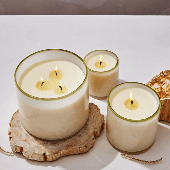 The Lafco - Feu De Bois Classic 6.5 Candle collection features three cream-colored candles in transparent glass jars, with hints of sandalwood. The largest candle sits on natural wood, creating a warm and cozy atmosphere on a light surface.