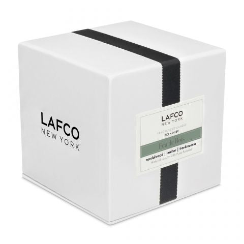 A Lafco - Feu De Bois Signature 15.5oz Candle is packaged in a white box with a vertical black ribbon, featuring sandalwood notes, a leather accord, and hints of frankincense.