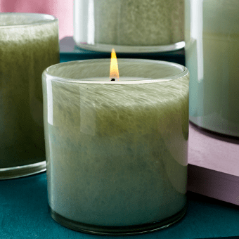 The Lafco Fresh Cut Gardenia Classic 6.5 Candle, elegantly placed in a glass holder with its gardenia aroma, is surrounded by unlit green candles on a pink and dark green surface for a harmonious setting.