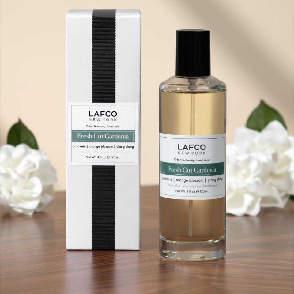 The Lafco - Fresh Cut Gardenia Room Mist is shown with its box on a wooden surface, accented by white gardenias and green leaves, bringing effective odor removal and a fresh floral scent to any room.