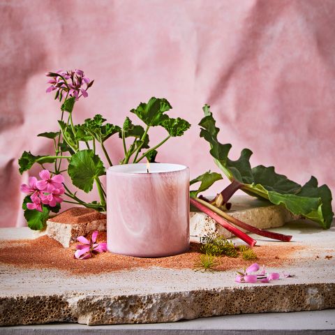 A Lafco - Geranium Bourbon Classic 15.5oz Candle is placed on stone with geranium bourbon flowers and green leaves. A nearby pile of red sand and rhubarb complements the display, set against a soft pink fabric backdrop.