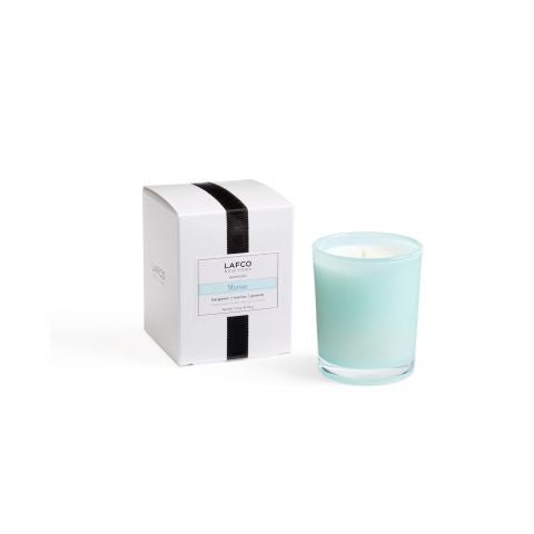 A Lafco Marine votive candle in light blue glass sits beside a white box with a black ribbon and LAFCO, Marine label. The unlit candle emits a subtle sea jasmine scent, while the box features matching light blue accents.