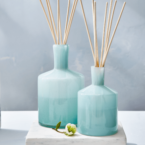 Two Lafco Marine 6oz Reed Diffusers, ideal for essential oil blends, stand with reed sticks on a marble surface. A small white flower with green leaves rests beside them, enhancing the tranquility of the soft, light blue background.