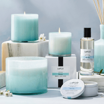 A Lafco Marine Classic 6.5 Candle in hand-blown glass is arranged with white flowers and marble blocks on a light gray surface, alongside other Marine Bergamot products like a tin, box, reed diffuser, and room spray.
