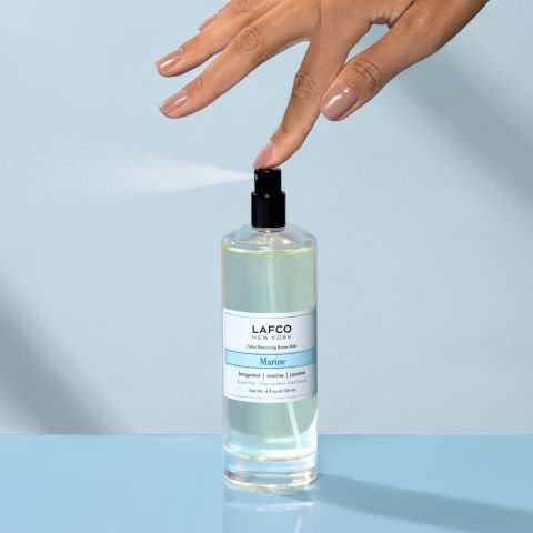 A manicured hand sprays Lafco - Marine Room Mist on a blue surface, releasing a fine marine-scented mist into the air.