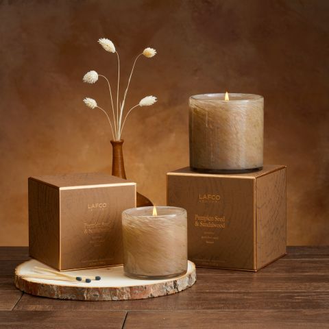 The Lafco Pumpkin Seed & Sandalwood Signature 6.5oz Candle glows on brown boxes atop wood, with matches nearby and dried white flowers in a vase against a warm brown backdrop, filling the air with patchouli and sandalwood notes.