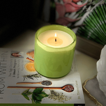 The Lafco Rosemary Eucalyptus Classic 6.5 Candle in a hand-blown glass vessel sits on a book with images of oranges, herbs, and spices, against a leafy backdrop and near a white decorative object.