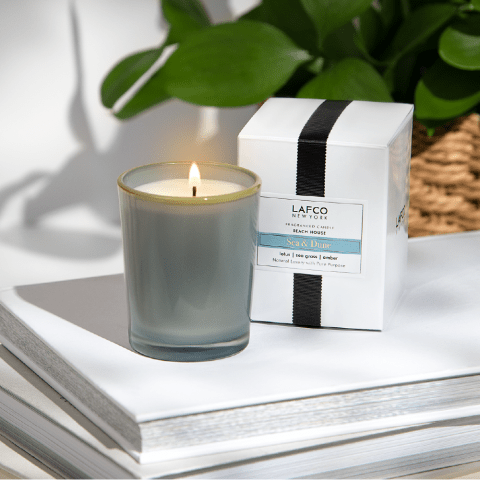 The Lafco Sea & Dune 1.9oz Votive Candle rests elegantly on stacked notebooks in a frosted glass holder, creating a serene atmosphere. A white box with a black stripe complements this BEACH HOUSE vibe, framed by greenery and enhanced by natural light casting soft shadows.