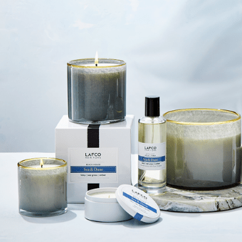 The Lafco Sea & Dune Classic collection features two lit candles in glass holders, an oil spray bottle, a labeled container, and a lotus-infused unlit candle on marble against a light blue background.