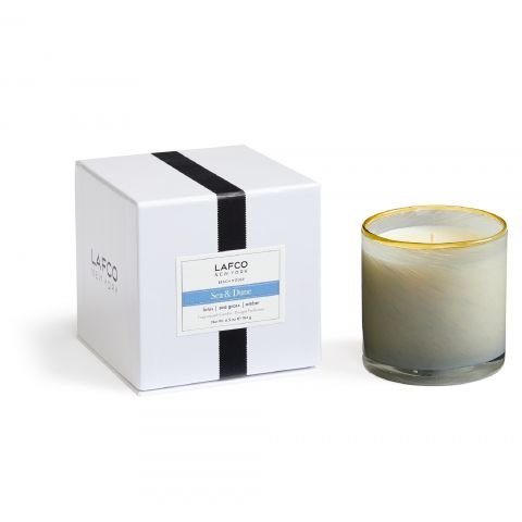 A Lafco Sea & Dune Classic 6.5 Candle features a white cube box with Lafco branding, a black stripe, and includes a glass holder candle with beige-gray marbling and gold trim, releasing gentle notes of amber and lotus.