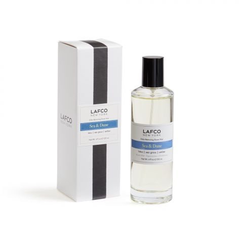 A bottle of Lafco Sea & Dune Room Mist, with essential oil-based fragrances, sits beside its box. The clear bottle holds a light liquid and features a blue and white label. The white box with a black stripe perfectly complements the elegant design of the label.