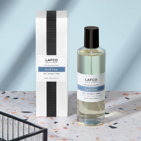 A bottle of Lafco - Sea & Dune Room Mist rests beside its box on a terrazzo surface. Scented with essential oil-based fragrances, it evokes a seaside escape. A light blue wall in the background casts shadows onto the scene.