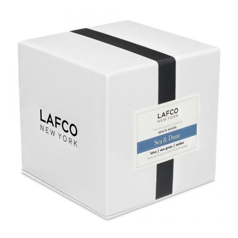 A white square box with a black vertical ribbon, labeled Lafco. The Sea & Dune Signature 15.5oz Candle features notes of lotus, sea grass, and amber for a luxurious essential oil experience.