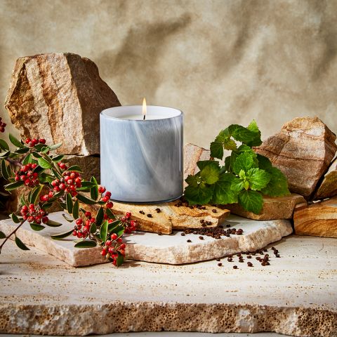 The Lafco - Timur Pepper Classic 15.5oz Candle features a lit wick in a marbled blue holder, surrounded by rocks, green leaves, and red berries on a stone surface with an earthy textured background.