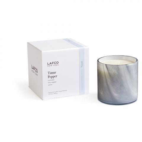 A white box labeled Lafco - Timur Pepper Classic 15.5oz Candle rests next to a marbled grey candle in a glass holder, with light blue accents on the box subtly reflecting the exotic black pepper scent as the candle peeks out.